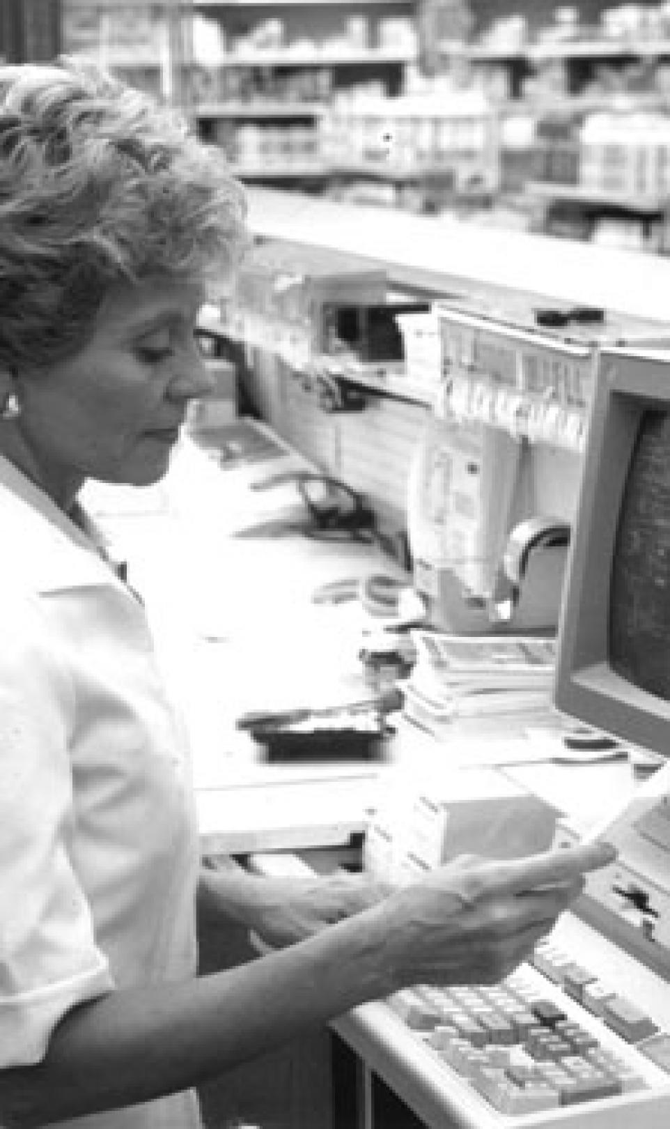 Black and white photograph showing a female employee, operating one of the first Intercom pharmacy computers behind the till of a Walgreens store