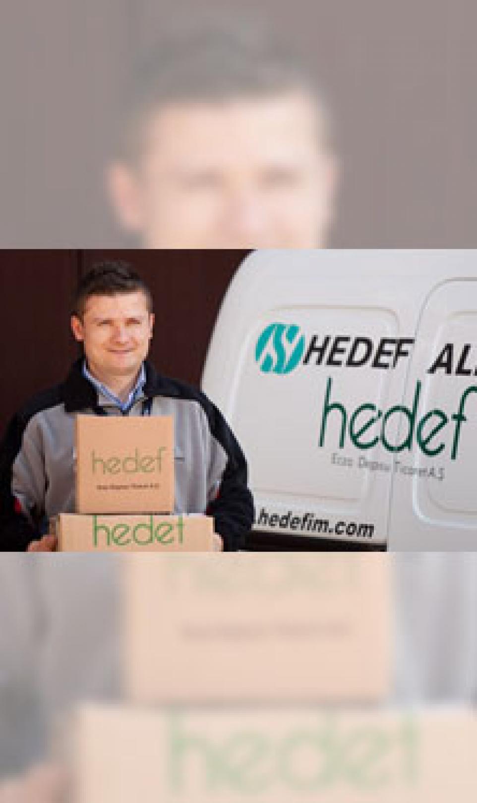 A man carries Hedef Alliance branded cardboard boxes next to a Hedef Alliance white van